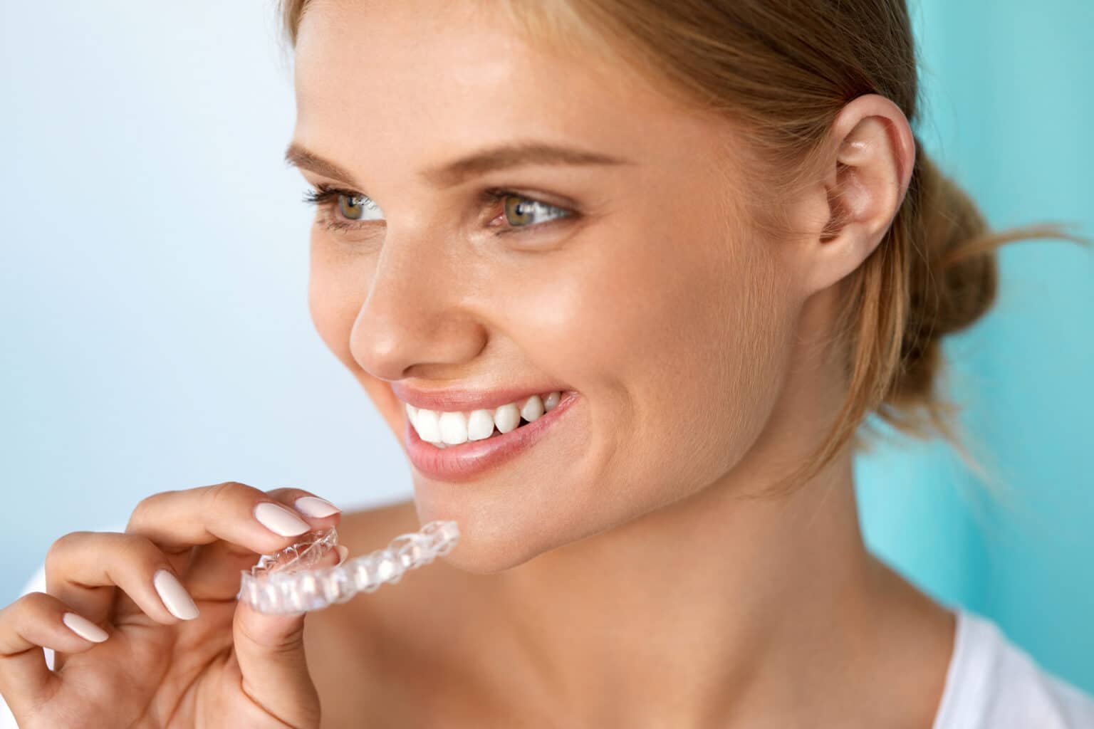 a blonde woman with straight white teeth holding her invisalign clear aligners near her face